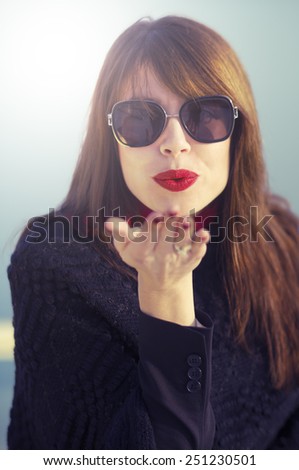 Beautiful elegant young woman wearing sunglasses and red lipstick blowing a kiss.