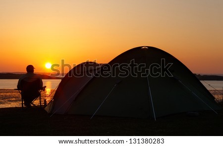 sun sets on the horizon behind a camper sitting relaxing and enjoying his camping holiday creating a silhouette of the pitched tent with him sitting beside