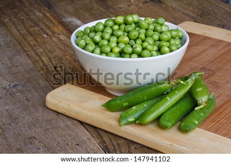 White Bowl of fresh garden peas recently taken from pea pod in a rustic kitchen setting with the pods in the foreground