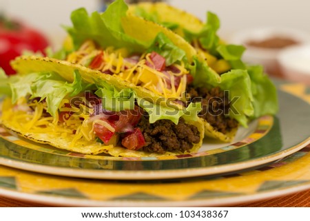 Beef Tacos - Mexican minced beef hard-shell tacos with salsa and cheese on a colourful plate.  Refried beans and sour cream on background.
