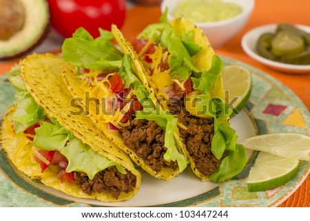 Beef Tacos - Mexican minced beef hard-shell tacos with salsa, cheese and lime wedges on a colourful plate. Guacamole and pickled jalapenos chilies on background.