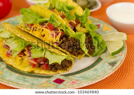 Beef Tacos - Mexican minced beef hard-shell tacos with salsa, cheese and lime wedges on a colourful plate. Sour cream and pickled jalapenos chilies on background.