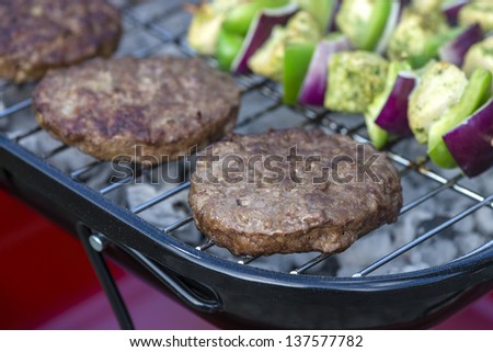 Burgers on BBQ - Patties being grilled on a portable barbeque outdoors. Chicken kebabs on background.