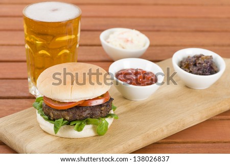 Burger & Beer -  Burger in a white bun with summer leaf and tomato served with ketchup caramelised onions, coleslaw and beer. Rustic outdoors setting.