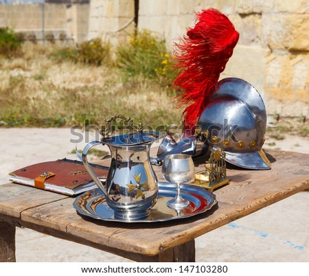 silver jug ??with a cup and a medieval helmet with a red pen on the wooden table