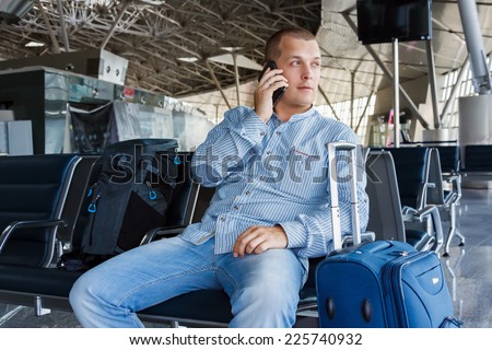 handsome young man with blond short hair talking on the phone, sitting on a chair with things at the airport waiting for his flight