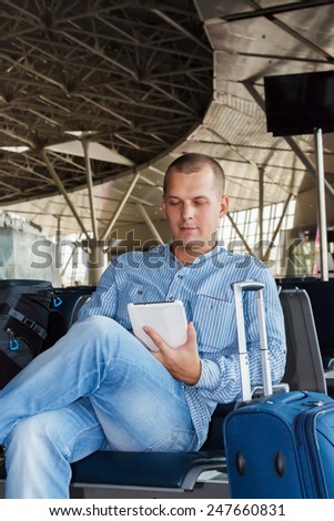 handsome young man with short hair working on the tablet, sitting on a chair with things at the airport waiting for his flight