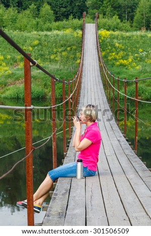 blonde girl with short haircut drinking tea from a thermos on Pedestrian suspension bridge of steel and wood over the river, summer in Russia