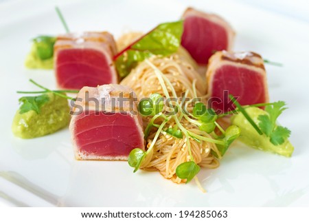 Grilled tuna chunks served with rice noodles and fresh green herbs on white plate. See more food shots