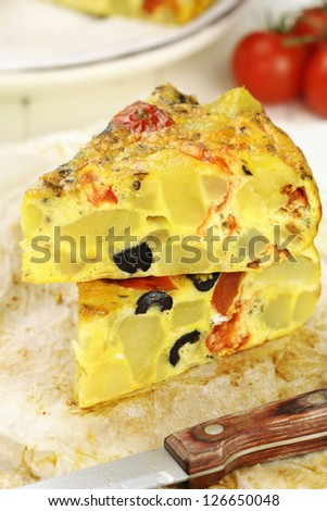 Slices of Spanish potato tortilla with black olives and tomatoes on a baking paper with a knife