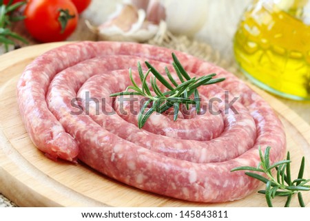 Raw Italian long pork sausage - LUGANEGA - on a wooden cutting board with olive oil and vegetables at the background ready to be cooked