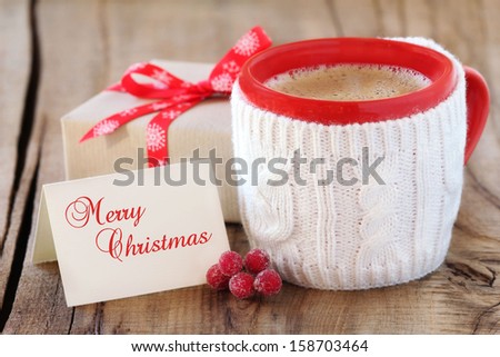 Cozy Rustic Christmas Setting - Red Mug Of Black Coffee In A White Knitted Cup Holder With A Wrapped Gift And A Greeting Card
