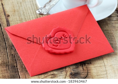 Love letter in a red envelope sealed with a red heart shaped sugar heart on a rustic wooden table with a white cup of coffee at the background