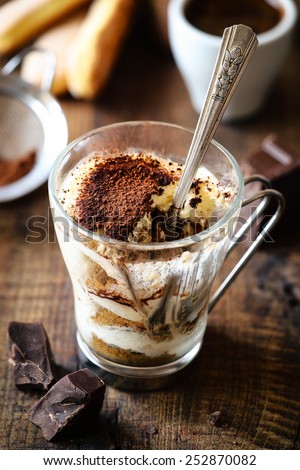 Half eaten Italian tiramisu dessert in a glass cup with a spoon with a cup of espresso coffee at the background, savoiardi and dark chocolate