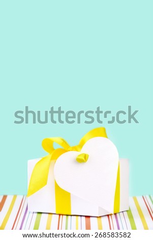 White gift box tied with a yellow satin ribbon with a heart shaped greeting card attached against turquoise background with copy space for Mother\'s Day, Easter, Birthday, Father\'s Day or any occasion