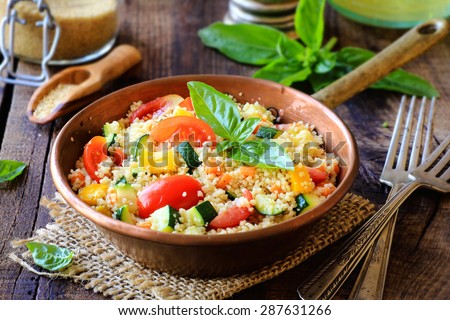 Delicious homemade vegetarian couscous with tomatoes, carrots, zucchini, yellow bell pepper and fresh basil on a dark rustic wooden kitchen table