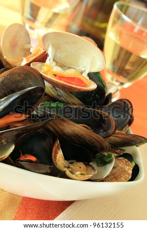 Fresh mussels and clams cooked in white wine with parsley, served in a white bowl with glasses of white wine at the background