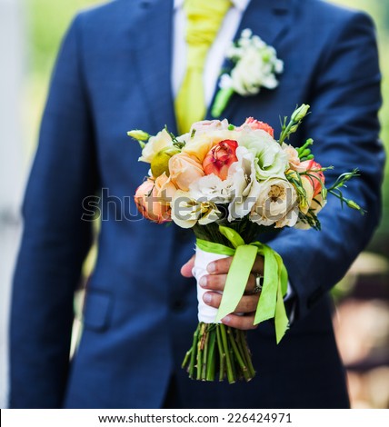 Beautiful wedding bouquet in hands of the groom. Gift to the bride. Roses, peonies taped and lace.Blue classic designer suit with a yellow tie and buttonhole.