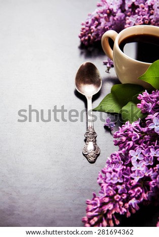Black coffee in a cup, sugar on a spoon and fresh lilac flowers on black background
