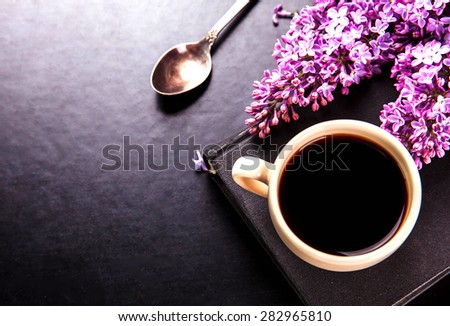 Black coffee in a cup, book, a spoon and fresh lilac flowers on black background