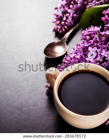 Black coffee in a cup, sugar on a spoon and fresh lilac flowers on black background