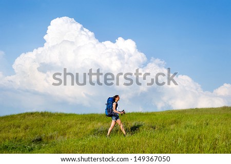 Smiling hiking young woman with backpack and trekking poles walking in a green meadow