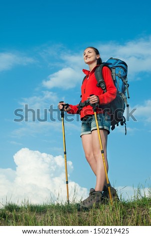 Smiling hiking young woman with backpack and trekking poles standing on a green meadow