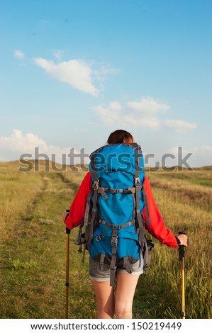 Hiking young woman with backpack and trekking poles walking in a green meadow