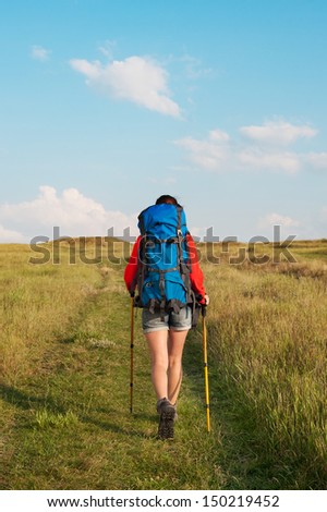 Hiking young woman with backpack and trekking poles walking in a green meadow