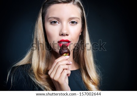 Beautiful young woman in a black dress holding delicious strawberry covered with black chocolate, isolated on a black background