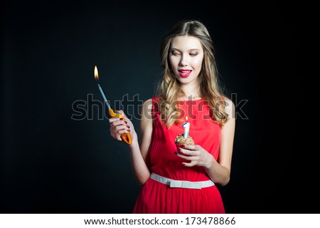 Portrait of beautiful young in a red dress holding birthday cupcake with one candle and gasoline lighter with flame on dark background