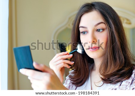 Portrait of beautiful young woman looking at the mirror putting on makeup holding a makeup brush