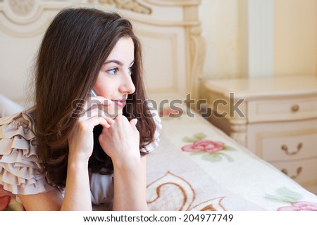 Portrait of a smiling young woman talking mobile phone lying on the bed in her bedroom