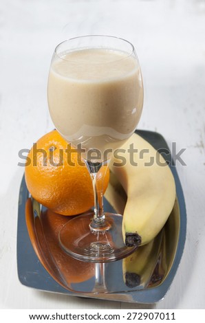 Smoothies of orange and banana in a glass on a    high leg on a metal tray . Bananas and oranges near. on a wooden white table.
