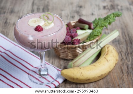 Smoothie of banana, frozen raspberries and rhubarb with yogurt in a glass. Bananas, frozen raspberries and rhubarb in the background on a wooden table.