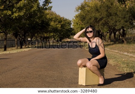 Brunette waiting for a lift sitting on suitcase roadside with hand in hair