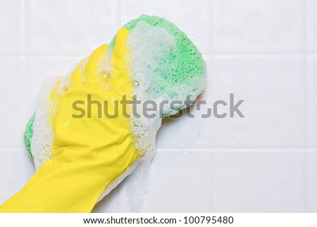 A yellow rubber gloved hand washing down a tile wall with a  soapy green microfiber sponge.