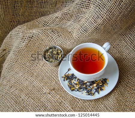 A cup of tea with loose tea in a container on a burlap surface