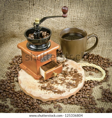 A cup of coffee grinder, cup of coffee and beans on wooden board on burlap