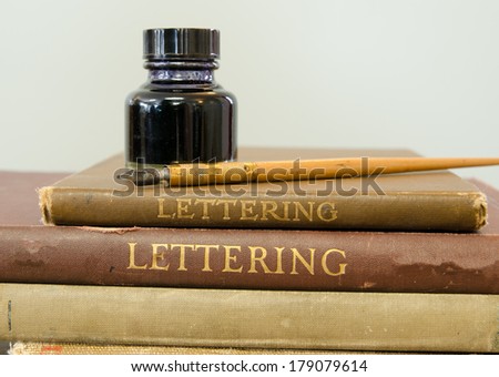 An old calligraphy pen and bottle of ink on some old lettering books.