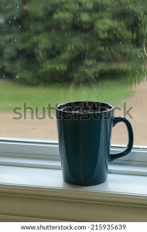 Steaming mug of coffee on a window sill with raindrops on window