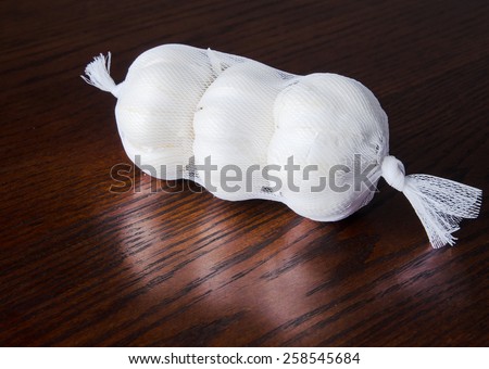 Three garlic heads packaged in a mesh bag on a polished rich wood surface with a reflection