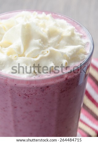 A pink berry smoothie in a frosty glass with whipped cream topping and a striped tea towel on a wood grain backdrop