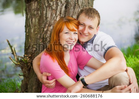 Portrait of happy couple in love. Man and woman sitting outdoors near big tree and river and looking at camera. Smiling family of young people.