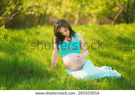 Young happy woman in expectation of baby. Female hands holding pregnant belly. Beautiful mother enjoying nature. Pregnant woman sitting on grass outside in summer park. Horizontal color photo.