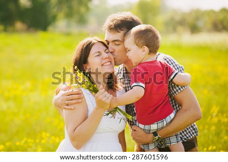 Happy family of three kissing outside. Mother in expectation of baby. Smiling faces of woman, man and child. Baby playing outdoors with parents. Togetherness. Man and woman hugging in sunset time.