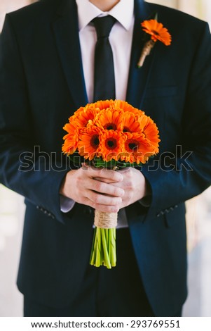 Groom in blue suit holding wedding bouquet  of flowers in hands waiting for his bride. Boutonniere and bridal bouquet of orange gerbera daisies.