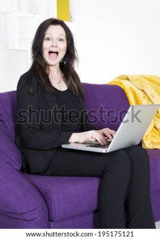woman in black suit sitting on purple couch with a laptop looking very happy