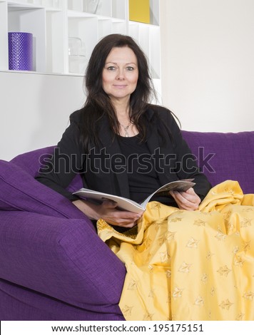 woman in a black suit sitting on purple couch with a yellow blanket and reading a magazine