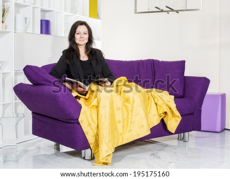 woman in a black suit sitting on purple couch with a yellow blanket and reading a magazine